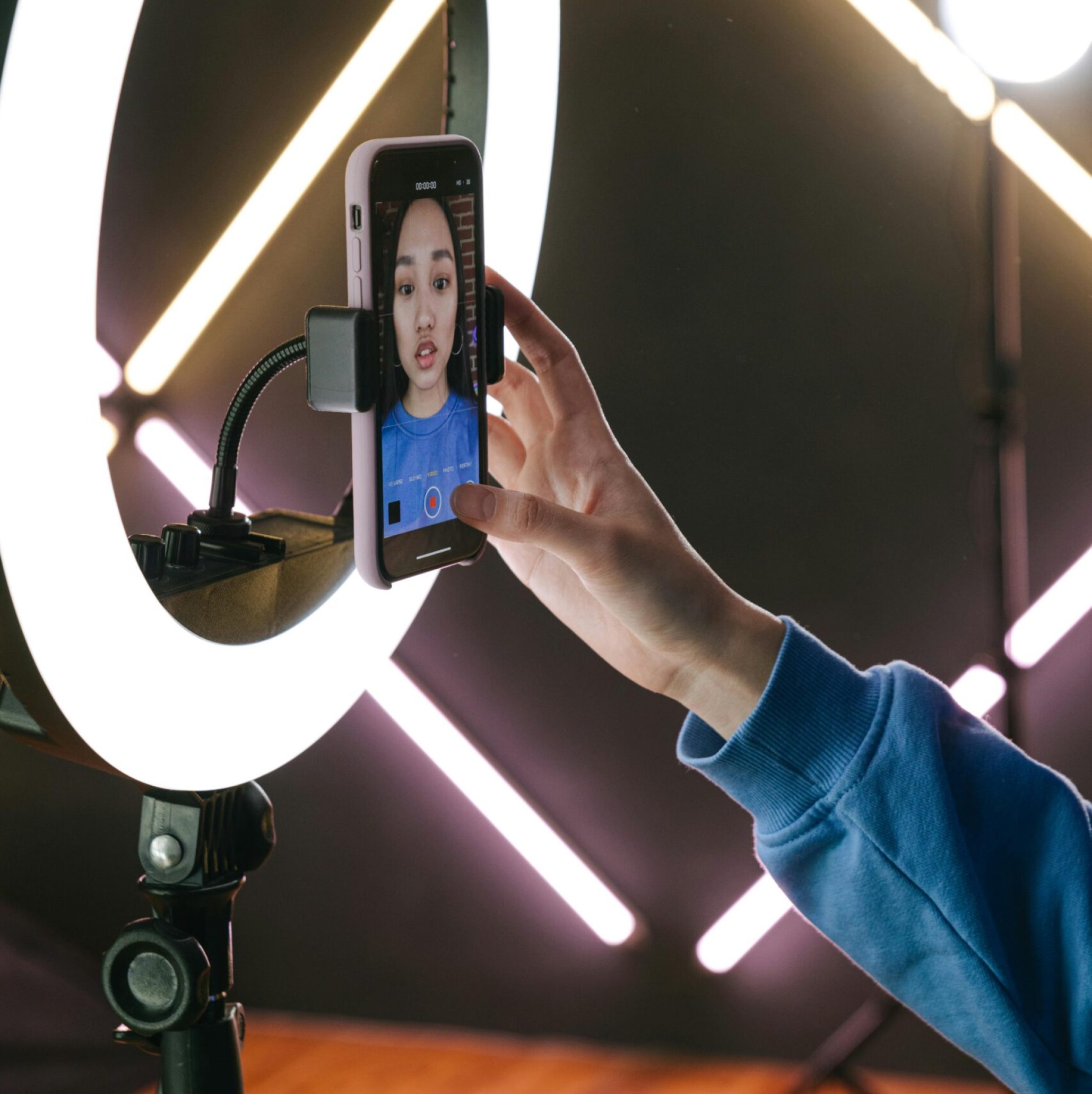 Asian woman using a smartphone and ring light for video streaming indoors, showcasing modern technology.